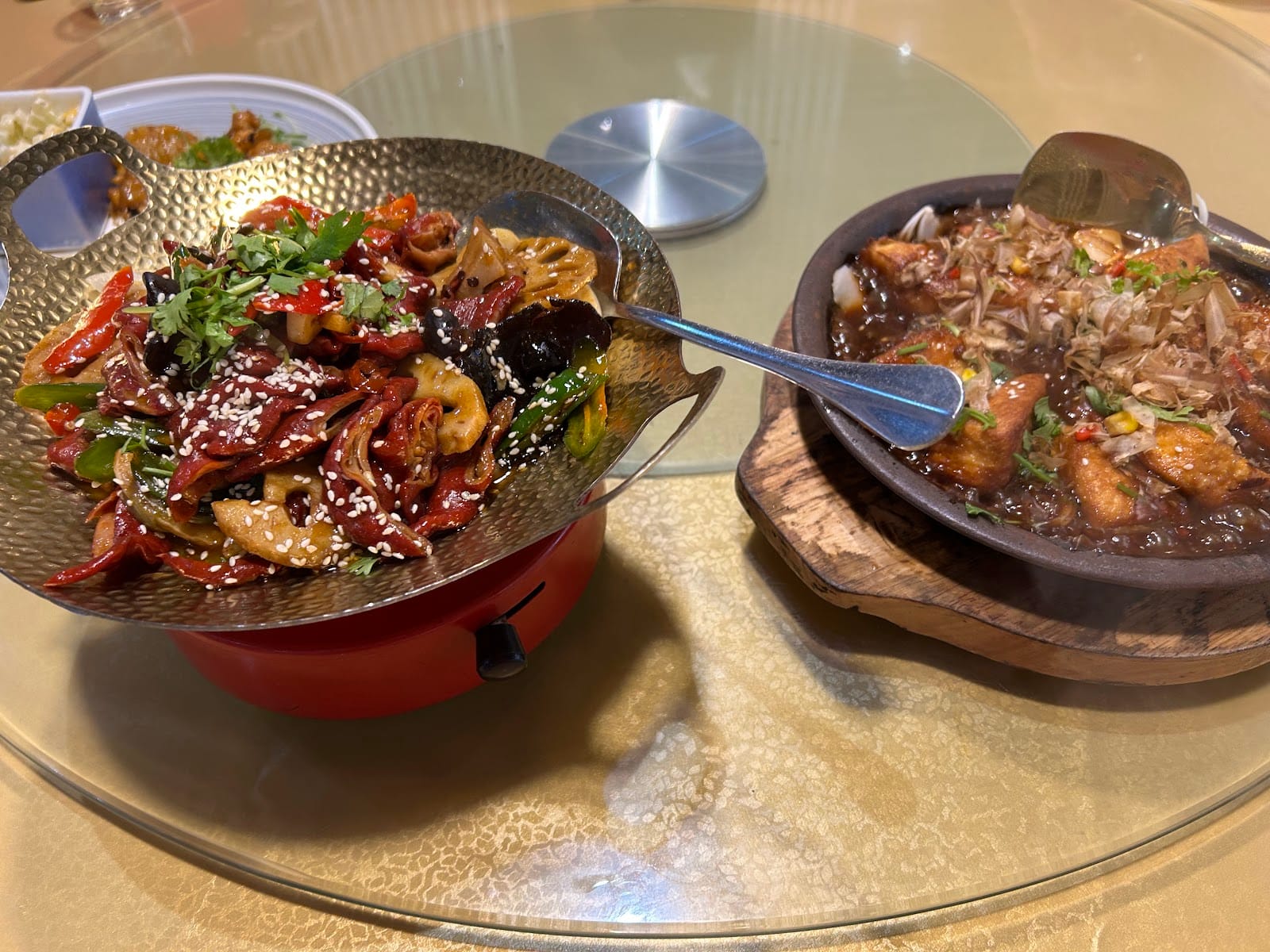 A hammered-metal dry pot of lotus root, wood ear and chilli over a burner, beside a stone pot of tofu under bonito flakes