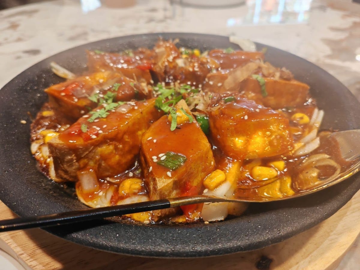 Close-up of tofu in a glossy sauce with corn and spring onion in a dark stone pot